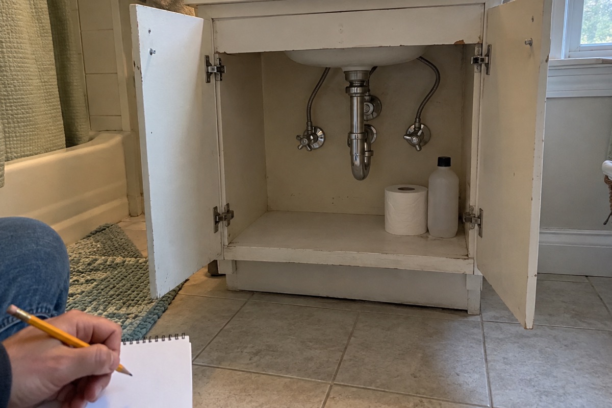 A bathroom vanity with its doors open shows braided supply lines and chrome shutoff valves, while a person seated on the floor writes on a notepad.