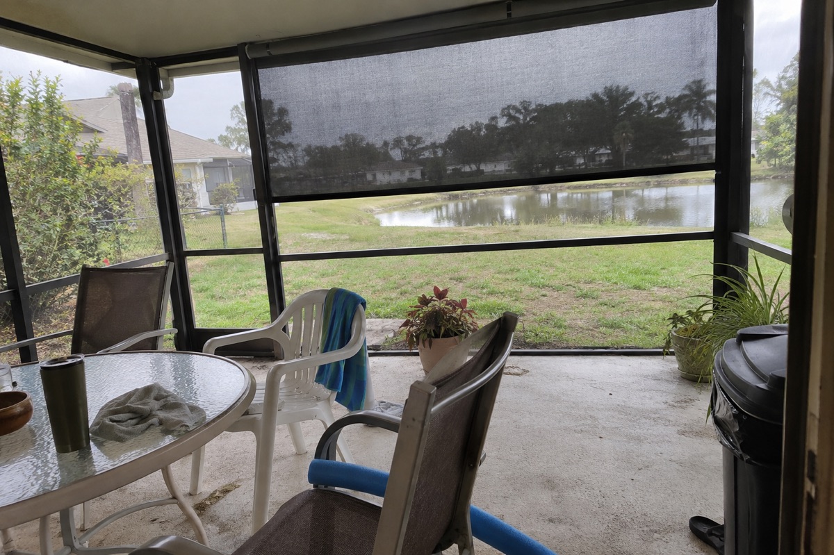 A half-lowered motorized mesh screen hangs across one side of an ordinary screened lanai with mismatched chairs.