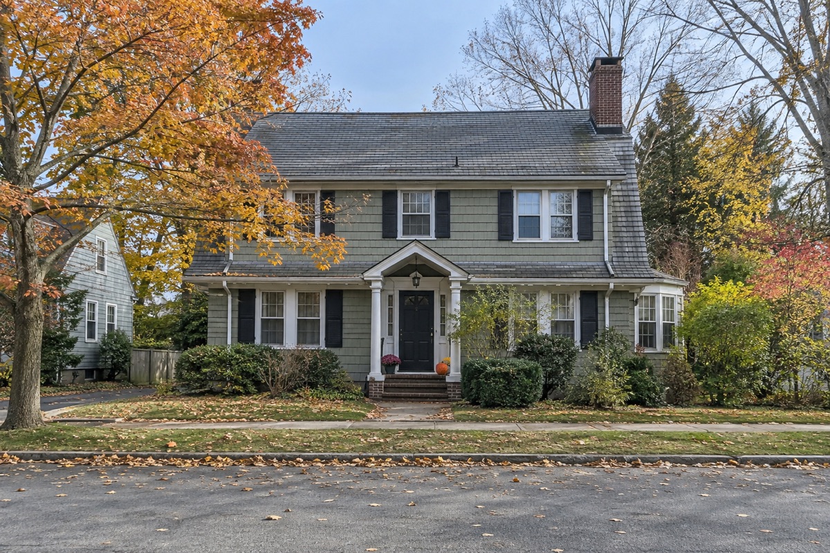 An older two-story home with a gray slate roof and brick chimney sits along an autumn street.