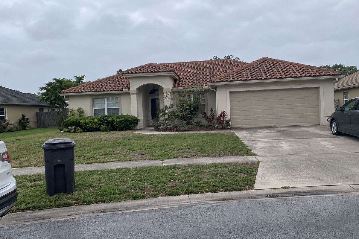 A one-story Florida stucco home with a terracotta tile roof is seen across a street beside a curbside trash bin.