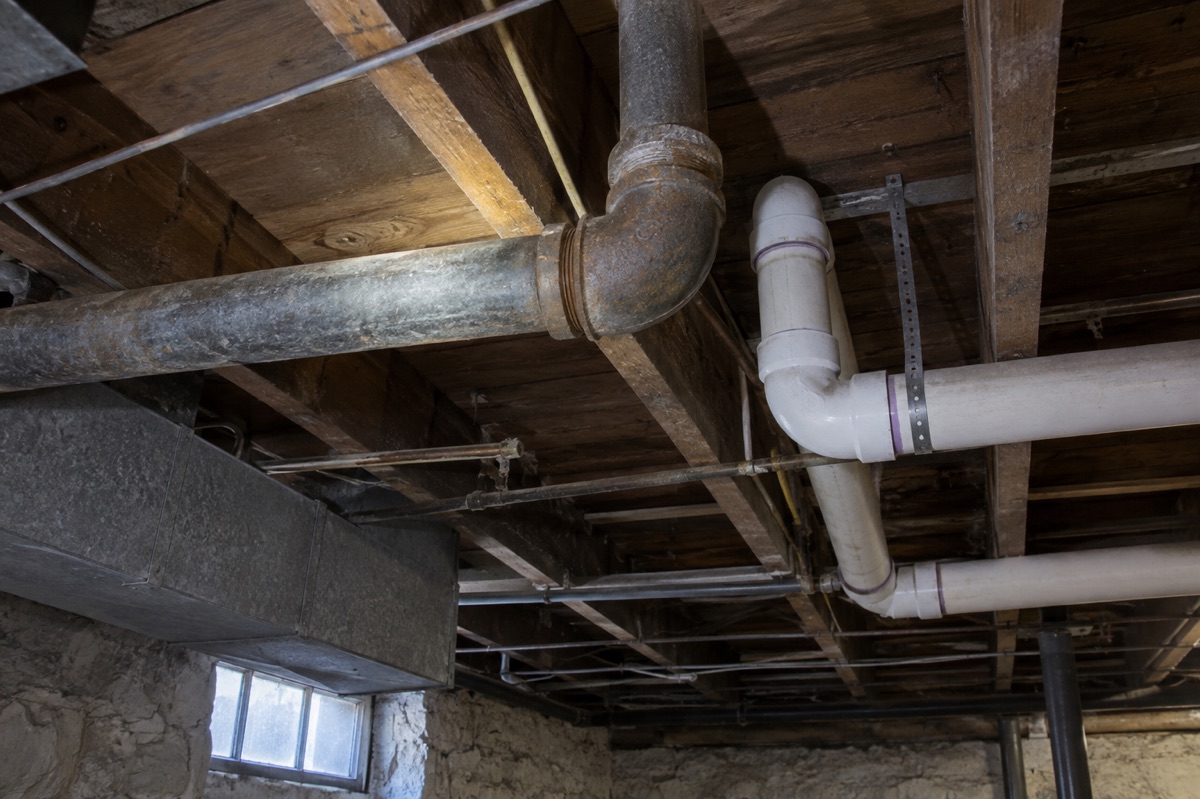 Older metal and newer plastic plumbing pipes run beneath an unfinished basement ceiling.