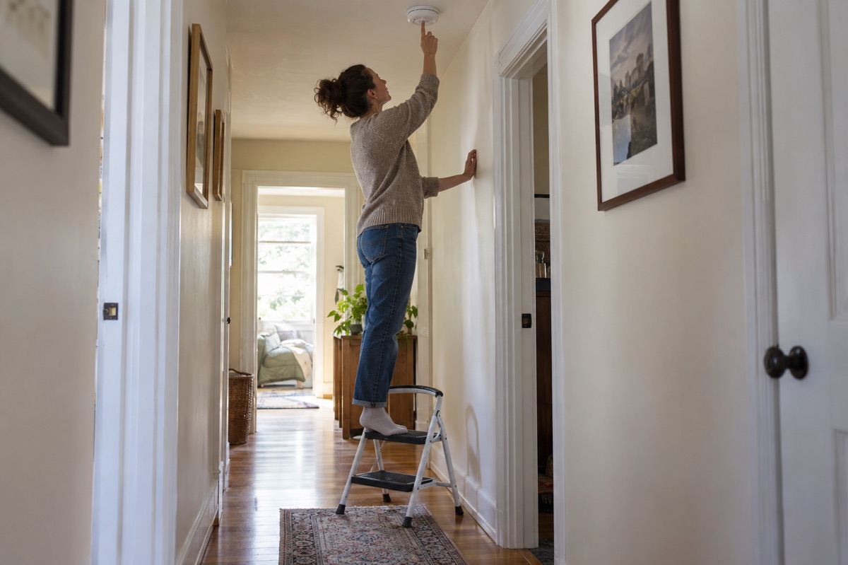 An adult on a sturdy two-step stool presses the test button on a hallway smoke alarm.