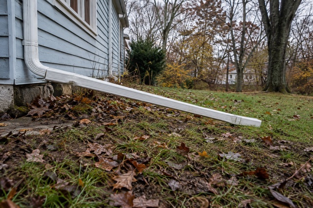 A downspout extension runs away from a home's foundation across a damp lawn.
