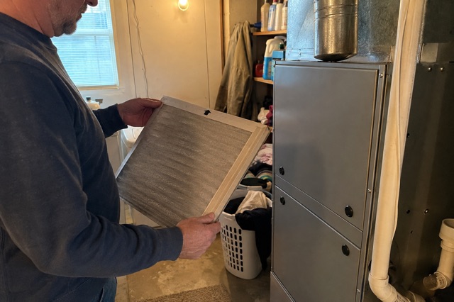 A homeowner checks a dusty HVAC filter beside a closed furnace cabinet in a lived-in utility room