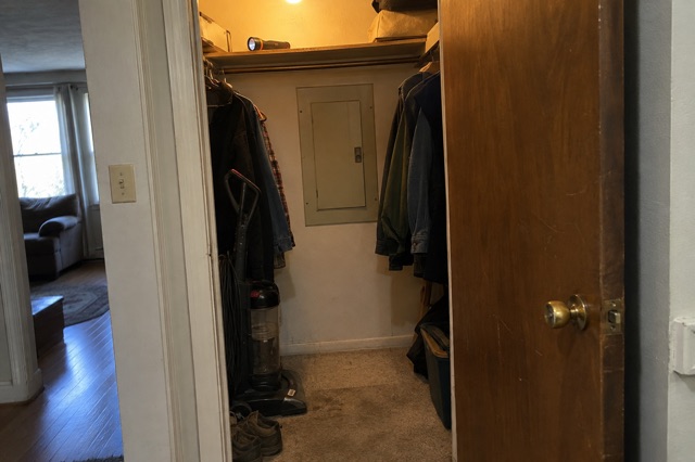 A hall closet with coats pushed to one side shows a closed beige breaker-panel door on the back wall and a clear patch of floor in front of it.