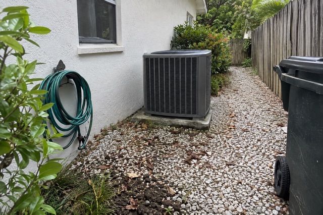A cleared gravel path along the side of a Florida block house leads to an outdoor air-conditioning unit, with trimmed shrubs pulled back and a coiled hose against the wall.