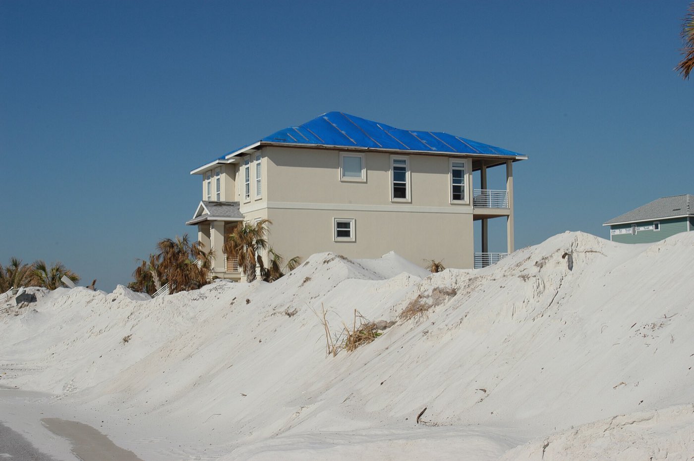 A two-story beach house with a blue tarp over its roof, behind mounds of storm-pushed sand